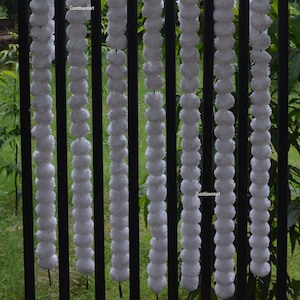 Op de afbeelding: Witte pompom-gordijn, een reeks witte, pluche ballen, hangend aan een zwart metalen hek. De gordijn is gemaakt van meerdere strengen pompoenen, elke streng is ongeveer 90 cm lang.
