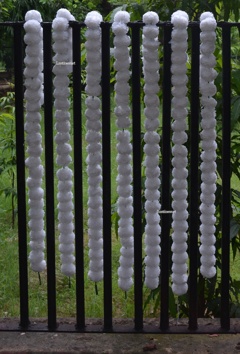 Op de afbeelding: Witte pompom-gordijn, een reeks witte, pluche ballen, hangend aan een zwart metalen hek. De gordijn is gemaakt van meerdere strengen pompoenen, elke streng is ongeveer 90 cm lang.