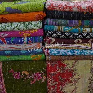 May include: A stack of colorful kantha quilts with various floral and geometric patterns. The quilts are made of recycled saris and are hand-stitched. The quilts are arranged in two stacks, one on the left and one on the right. The quilts are all different sizes and shapes. The quilts are all made of cotton and are very soft. The quilts are perfect for adding a touch of color and style to any room.
