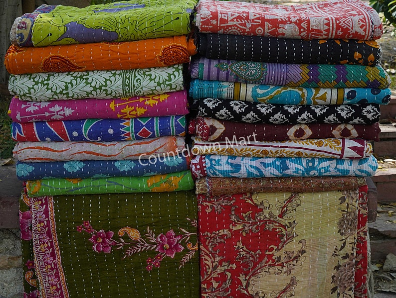 May include: A stack of colorful kantha quilts with various floral and geometric patterns. The quilts are made of recycled saris and are hand-stitched. The quilts are arranged in two stacks, one on the left and one on the right. The quilts are all different sizes and shapes. The quilts are all made of cotton and are very soft. The quilts are perfect for adding a touch of color and style to any room.