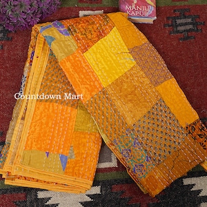 Peut inclure: Une courtepointe patchwork aux couleurs vives d'orange, de jaune et d'or. Elle présente divers motifs géométriques et textures, avec des coutures visibles. Le texte "Countdown Mart" est visible. Un livre et des fleurs violettes sont en arrière-plan.