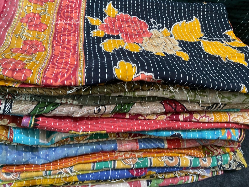 May include: A stack of colorful kantha quilts with floral patterns. The quilts are made of recycled saris and are hand-stitched. The top quilt is black with yellow and red floral designs.