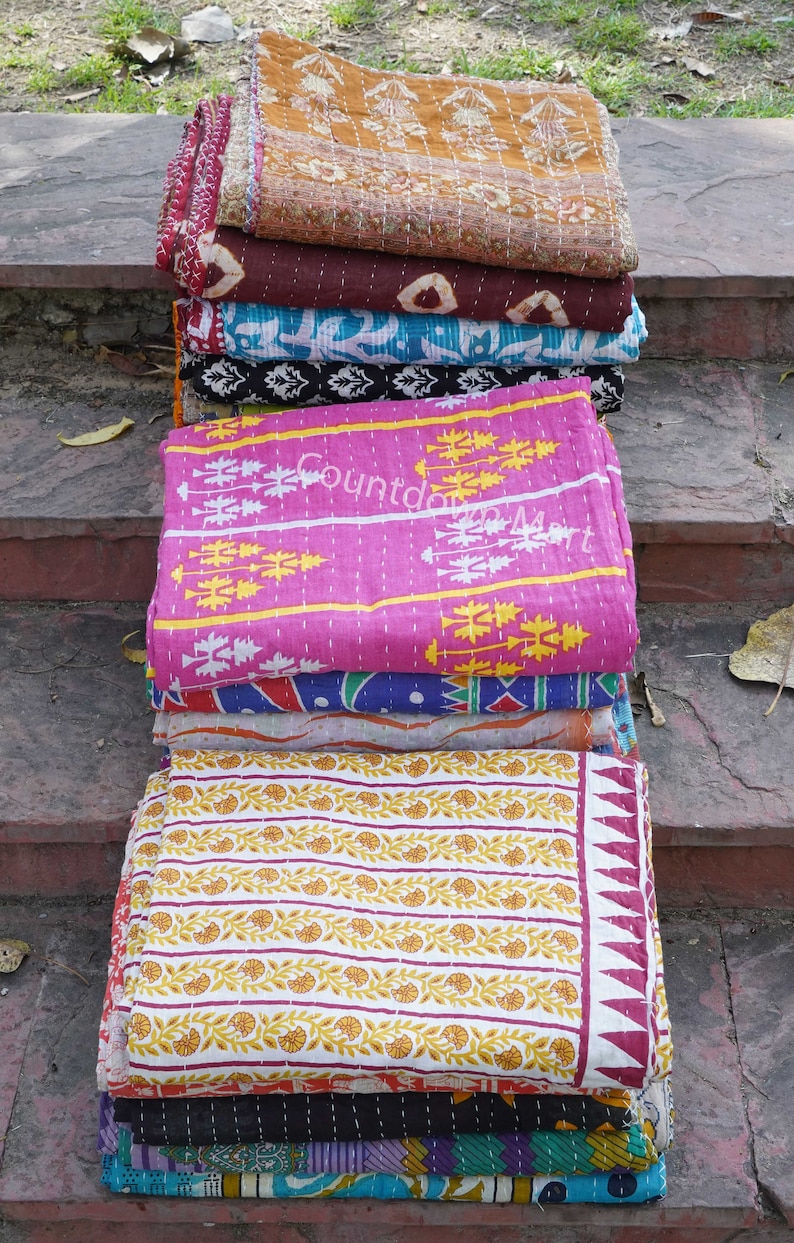 May include: A stack of colorful kantha quilts with various floral and geometric patterns. The quilts are made of cotton and are hand-stitched. The quilts are folded and stacked on top of each other, creating a visually appealing display.