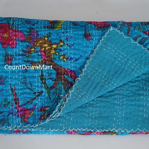 May include: A folded, turquoise quilt with a floral pattern in pink, red, and brown. The reverse side is a solid turquoise color. White stitching outlines the edges and patterns.