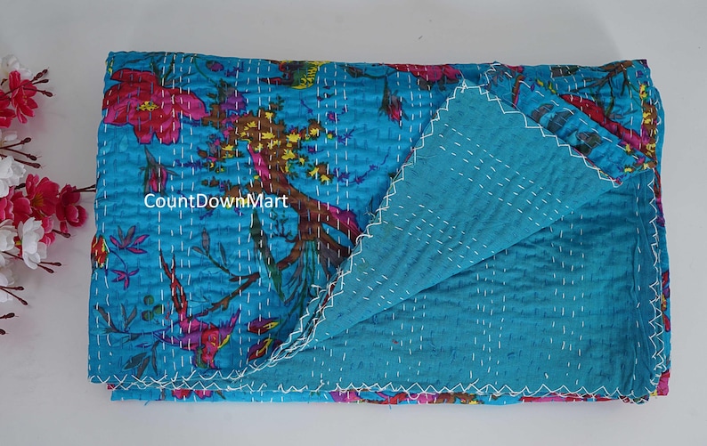 May include: A folded, turquoise quilt with a floral pattern in pink, red, and brown. The reverse side is a solid turquoise color. White stitching outlines the edges and patterns.