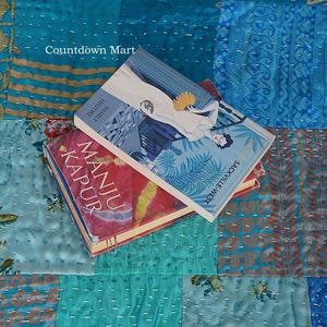 Könnte beinhalten: Zwei Bücher mit farbenfrohen Einbänden liegen auf einem Patchwork-Quilt. Das obere Buch hat einen blauen Einband mit einer Frau, die einen Fächer hält. Das untere Buch hat einen roten Einband mit dem Namen des Autors, Manju Kapur. Der Quilt besteht aus blauen und türkisfarbenen Stoffen.