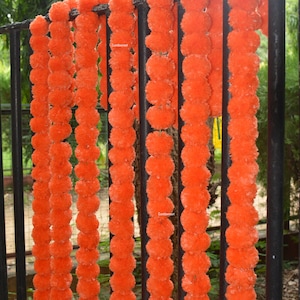 Op de afbeelding: Een close-up van een guirlande van oranje stoffen bloemen, hangend aan een metalen hek. De guirlande is gemaakt van veel kleine, ronde pompoenen en is perfect om een feestelijke touch toe te voegen aan elke gelegenheid.