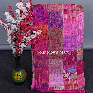 Könnte beinhalten: Eine lebendige Patchwork-Decke in Rosa-, Magenta- und Lilatönen. Die Decke weist eine Vielzahl von gemusterten Stoffen auf, die mit sichtbarem Faden zusammengenäht sind. Der Text "Countdown Mart" ist auf der Decke zu sehen. Im Hintergrund eine dekorative Vase mit Blumen.