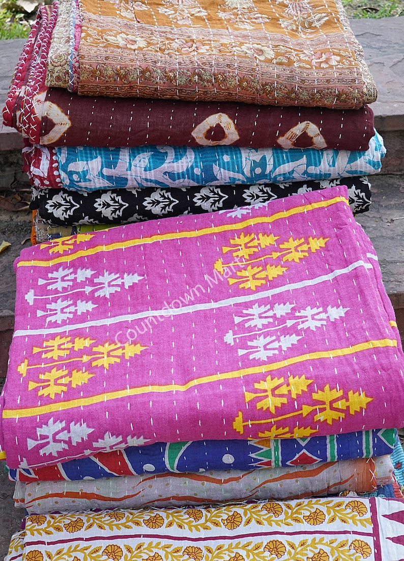 May include: A stack of colorful kantha quilts with various patterns, including floral, geometric, and paisley designs. The quilts are made of cotton and are hand-stitched. The top quilt is pink with white and yellow geometric patterns.