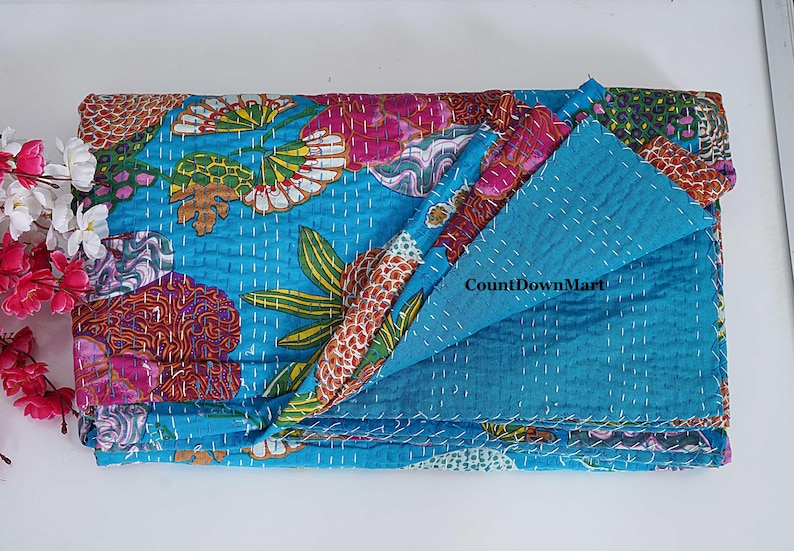 May include: A folded, turquoise quilt with a floral pattern in pink, orange, and green. The quilt is hand-stitched with white thread. The text "CountDownMart" is visible on the quilt. The quilt is folded on a white surface.
