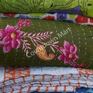 May include: A stack of kantha quilts in various colors and patterns, including green with floral designs, orange and white striped, and blue with small floral designs. The top quilt has the text "Countdown Mart" printed on it.
