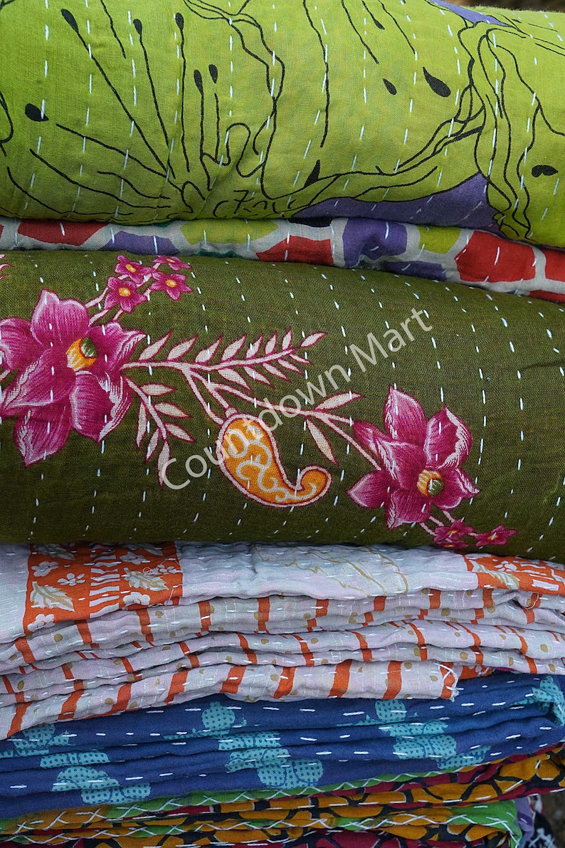 May include: A stack of kantha quilts in various colors and patterns, including green with floral designs, orange and white striped, and blue with small floral designs. The top quilt has the text "Countdown Mart" printed on it.