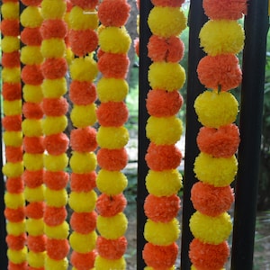 Op de afbeelding: Een guirlande van gele en oranje stoffen bloemen aan elkaar geregen. De guirlande hangt aan een zwart metalen hek.