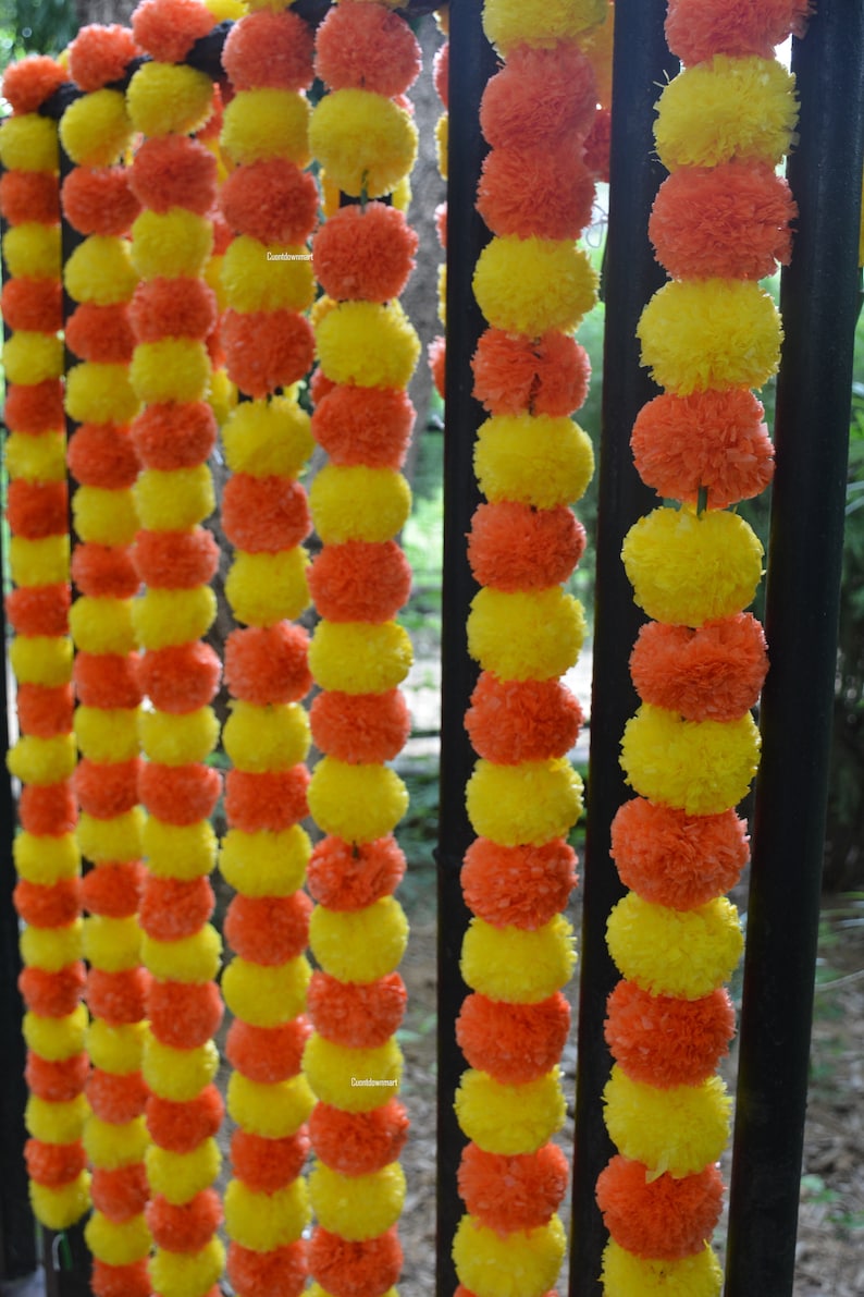 Op de afbeelding: Een guirlande van gele en oranje stoffen bloemen aan elkaar geregen. De guirlande hangt aan een zwart metalen hek.