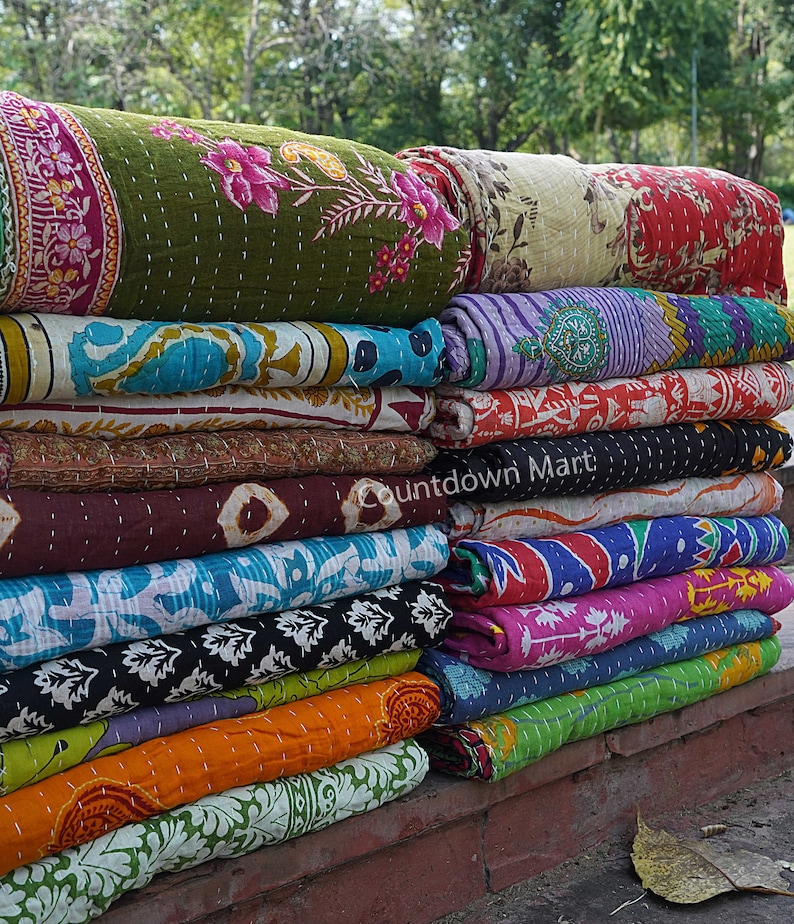 May include: A stack of colorful kantha quilts, a traditional Indian textile, with various floral and geometric patterns. The quilts are folded and stacked on top of each other, creating a visually appealing display.
