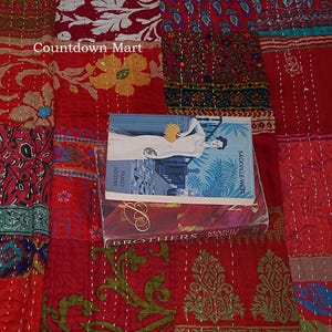 Könnte beinhalten: Ein roter Patchwork-Quilt mit Blumen- und Paisley-Mustern. Mehrere Bücher liegen darauf, darunter eines mit einer Frau auf dem Cover und dem Text "SACKVILLE-WEST". Der Quilt hat ein gesticktes Design.