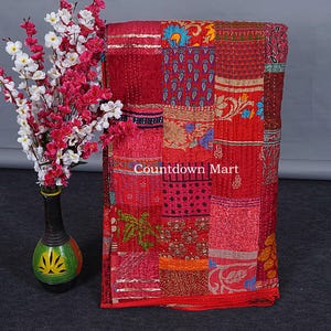 Könnte beinhalten: Ein Patchwork-Quilt in Rottönen mit verschiedenen Mustern und Texturen. Der Quilt besteht aus verschiedenen Stoffstücken, die zusammengenäht sind. Der Text "Countdown Mart" ist sichtbar. Links ist eine Vase mit Blumen.