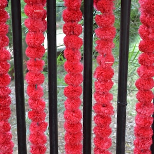 Op de afbeelding: Een close-up van een rode krans van kunstbloemen die aan een zwart metalen hek hangt. De krans is gemaakt van veel kleine, pluche bloemen.