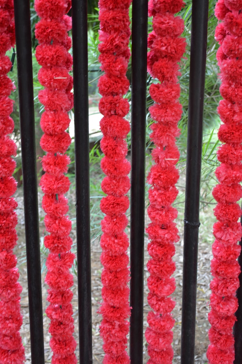 Op de afbeelding: Een close-up van een rode krans van kunstbloemen die aan een zwart metalen hek hangt. De krans is gemaakt van veel kleine, pluche bloemen.
