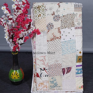 May include: A folded patchwork quilt with a variety of patterned fabric squares in white, cream, and various colors. The quilt features floral, geometric, and abstract designs. A vase with pink and white flowers is to the left.