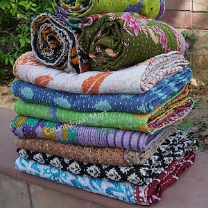 May include: A stack of colorful kantha quilts, a traditional Indian textile, with various floral and geometric patterns. The quilts are made of recycled saris and are hand-stitched with running stitch.  The quilts are folded and stacked on a stone surface.