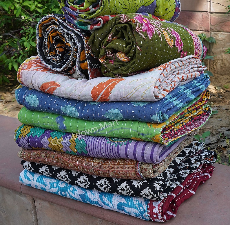 May include: A stack of colorful kantha quilts, a traditional Indian textile, with various floral and geometric patterns. The quilts are made of recycled saris and are hand-stitched with running stitch.  The quilts are folded and stacked on a stone surface.