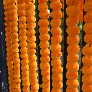 Könnte beinhalten: Nahaufnahme einer Girlande aus orangefarbenen Ringelblumen, die an einem schwarzen Metallzaun befestigt sind. Die Blumen sind in einem sich wiederholenden Muster angeordnet, was ein optisch ansprechendes und festliches Erscheinungsbild erzeugt.