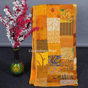 May include: A patchwork quilt in shades of yellow and gold, featuring a variety of patterned squares. The quilt has a stitched design and a border. A vase with pink and white flowers is on the left.