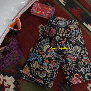 May include: Black floral quilt with colorful flower and paisley designs. The quilt is folded and displayed on a patterned rug. A book and a bundle of purple flowers are also visible.