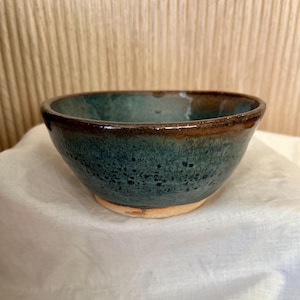 May include: A small, dark green ceramic bowl with a brown rim. The bowl has a speckled glaze and a slightly uneven surface.