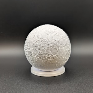May include: A white, spherical lamp designed to resemble the moon, with a textured surface showing craters and other lunar features. The lamp rests on a small, circular white base, creating a celestial decorative item.