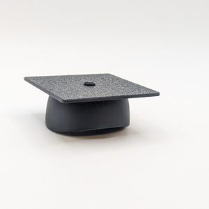 May include: A miniature black graduation cap with a textured square top and a rounded base. The cap is a solid black color and sits on a white background. The cap is a symbol of academic achievement.