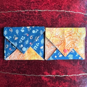 Peut inclure: Deux portefeuilles en tissu avec fermetures à bouton. L'un présente un motif floral bleu marine avec un accent jaune, et l'autre un motif jaune et orange avec un accent bleu. Les portefeuilles sont probablement faits à la main et adaptés aux cartes ou petits objets.