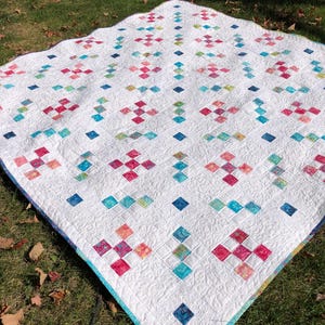 Peut inclure: Une courtepointe blanche avec un motif de patchwork composé de carrés de différentes couleurs, notamment rose, bleu, vert et jaune. La courtepointe est posée sur une surface herbeuse.