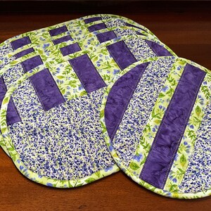 May include: Set of six purple and green quilted placemats with a floral pattern. The placemats are rectangular and have rounded corners. There is also a matching round placemat.