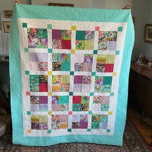 May include: A patchwork quilt with a teal border and a white background. The quilt is made up of squares of different fabrics with floral patterns in shades of pink, purple, green, and yellow.