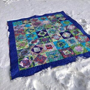 Puede incluir: Un edredón colorido con un borde azul descansa sobre la nieve. El diseño de patchwork presenta varios patrones y colores, incluyendo azul, verde, morado y rojo. El edredón rectangular parece hecho a mano.