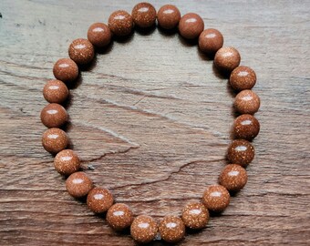 May include: A goldstone bracelet featuring spherical, sparkling beads. The beads have a speckled texture and a warm, brown hue. The bracelet is presented on a wooden surface, highlighting the jewellery's texture and colour.