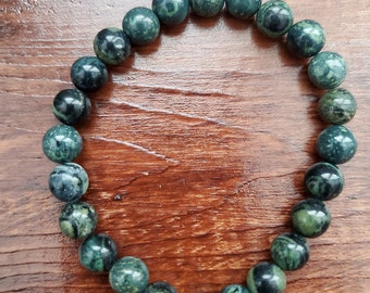May include: A dark green beaded bracelet with a mix of lighter green and black accents. The round beads are strung together, forming a complete circle. The bracelet is displayed on a wooden surface.