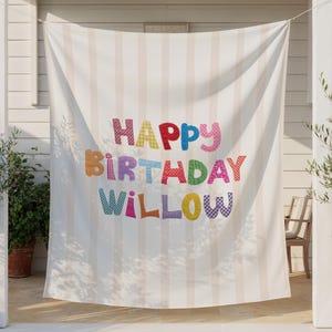 May include: A large, rectangular fabric banner featuring vertical light beige stripes. The text "HAPPY BIRTHDAY WILLOW" is printed in the center in large, multi-colored, patterned block letters including red, blue, green, yellow, and purple.