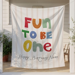 May include: A white fabric banner with vertical beige stripes featuring the colorful text "FUN TO BE ONE" in various patterns including polka dots and gingham. Below the main text, the words "Happy Birthday Noah" are written in black script.