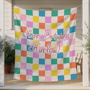 May include: A colorful checkered banner with the words "Happy Birthday Harlow!" in pink script. The squares are in shades of yellow, orange, pink, green, blue, and white. The banner is hanging outside.