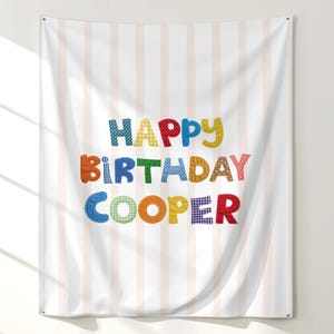 May include: A rectangular fabric wall tapestry featuring vertical light pink and white stripes. The text "HAPPY BIRTHDAY COOPER" is printed in the center using large, multi-colored letters with various patterns including polka dots and gingham.