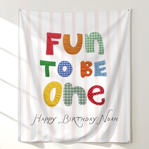 May include: A rectangular fabric wall hanging featuring a light pink and white vertical striped background. The text "FUN TO BE one" is printed in large, multi-colored, patterned letters. Below, the text "HAPPY BIRTHDAY NOAH" is in black.