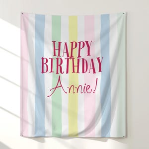 May include: A vertical striped wall hanging with pastel colors of pink, blue, yellow, and green. The words "HAPPY BIRTHDAY Annie!" are printed in a bold, pink font, making it a festive birthday decoration.