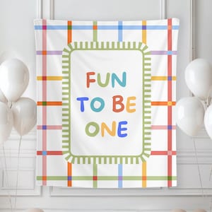 May include: A white banner with a colorful plaid pattern and the words "FUN TO BE ONE" in a rectangular frame. White balloons and beige balloons are arranged around the banner, creating a festive atmosphere.