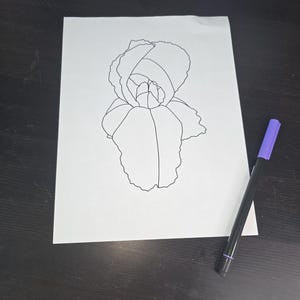 May include: A black and white line drawing of a flower on a white sheet of paper. A purple and black marker is placed next to the drawing. The flower drawing is in the center of the paper.