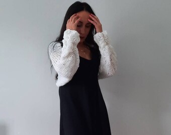 May include: A white crocheted shrug with long sleeves and a loose fit. The shrug is worn over a black dress.