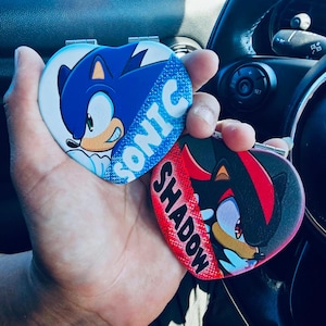 Könnte beinhalten: Zwei herzförmige Kompaktspiegel mit Sonic the Hedgehog und Shadow the Hedgehog. Der Sonic-Spiegel ist blau und weiß mit dem Wort "SONIC". Der Shadow-Spiegel ist schwarz, rot und weiß mit dem Wort "SHADOW".