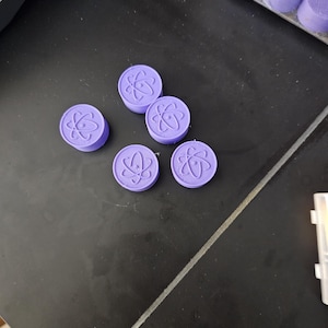 May include: Several small, round, lavender-colored objects with a raised atom design are scattered on a dark surface. The objects appear to be small, possibly decorative or functional, with a consistent design.